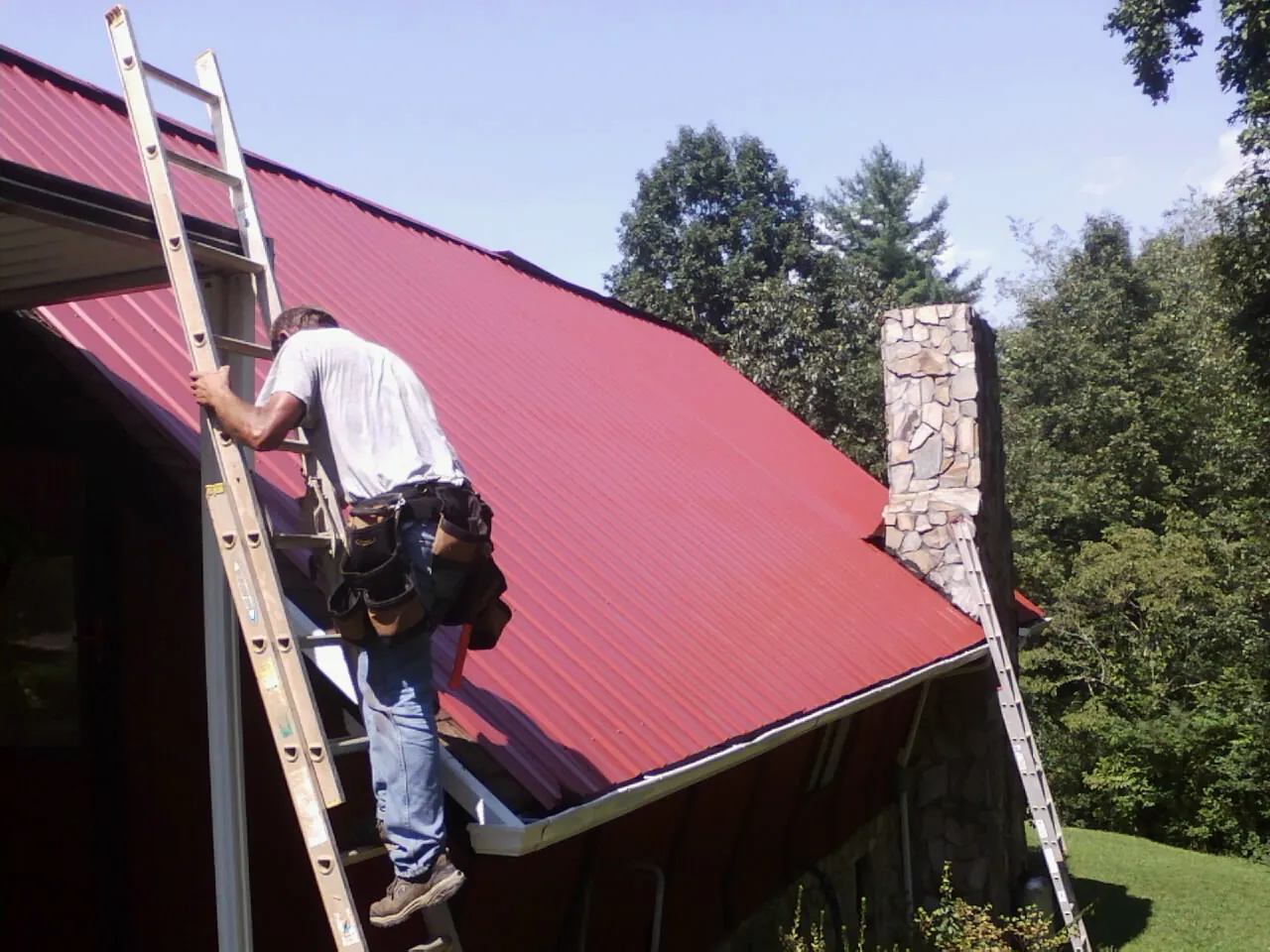 Roofer on a ladder beside a red metal roof and stone chimney — Soffit & Fascia Repair by Roofing Brothers in Vilas
